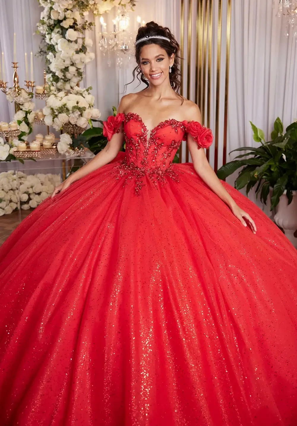 A woman in a glamorous red ball gown with off-shoulder sleeves and floral embellishments, standing in an elegant setting adorned with white flowers and greenery.