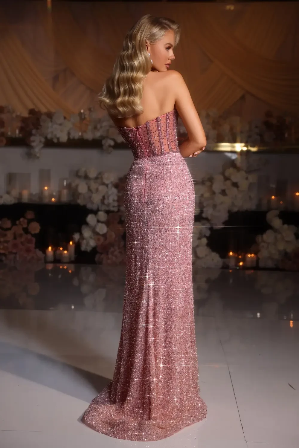 A woman in a sparkling pink gown, viewed from the back, with long hair styled in loose waves, standing in a dimly lit room adorned with floral decorations.